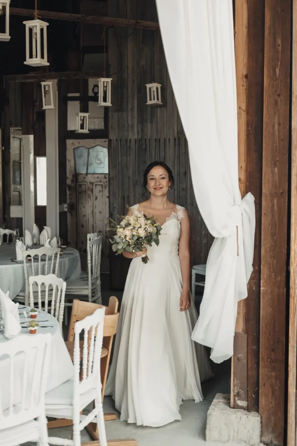Die Braut betritt mit einem strahlenden Lächeln die rustikale Hochzeitslocation Lindenhof in Mötzingen. Sanft fallende Stoffbahnen und hängende Laternen sorgen für eine warme, romantische Atmosphäre.
