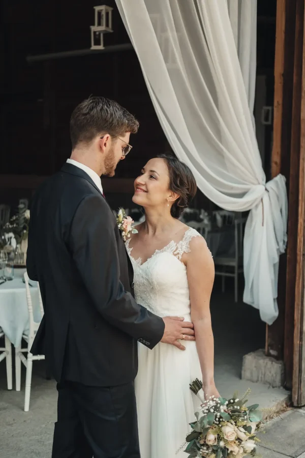 Hochzeitspaar vor einer rustikalen Eventlocation, umgeben von weißen Vorhängen – romantischer Moment voller Vorfreude.