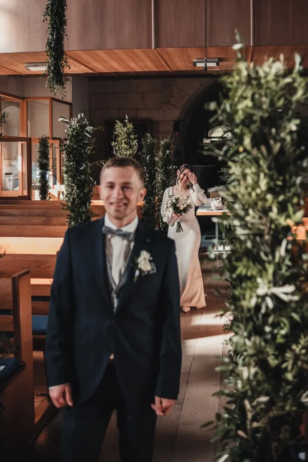 Emotionale First-Look-Szene in der Martinskirche Sielmingen: Der Bräutigam steht gespannt vorne, während die Braut mit glücklichem Lächeln und Blumenstrauß auf ihn zukommt. Mediterrane Dekoration mit Olivenzweigen rahmt die Szene ein.