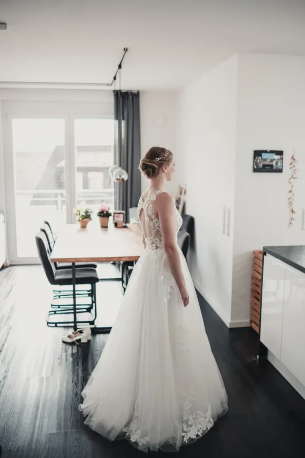 Rückansicht der Braut, die ihr prachtvolles, mit floraler Spitze verziertes Kleid präsentiert – ein strahlender Moment der Ruhe vor der Trauung.