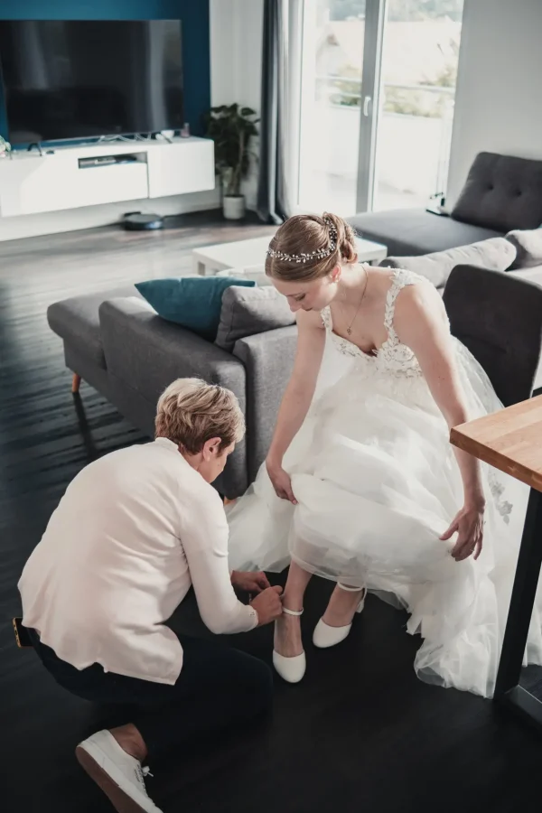 Die Braut, unterstützt von einer helfenden Hand, zieht ihre eleganten weißen Hochzeitsschuhe an – ein Moment voller Anmut und Eleganz.