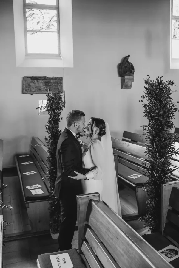 Das Brautpaar schreitet gemeinsam durch den mit Olivenzweigen geschmückten Mittelgang der Martinskirche. Der Bräutigam blickt seine Braut voller Liebe an, während sie mit strahlendem Lächeln ihren Brautstrauß hält.