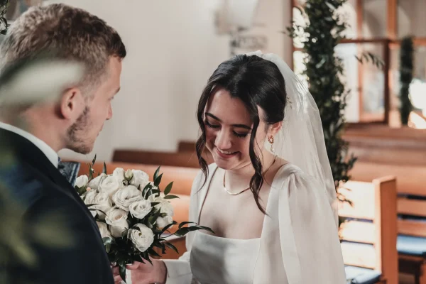 Innige Nahaufnahme: Der Bräutigam hält den Brautstrauß, während die Braut mit einem sanften Lächeln zu Boden blickt. Die goldene Lichtstimmung verleiht dem Moment besondere Wärme.