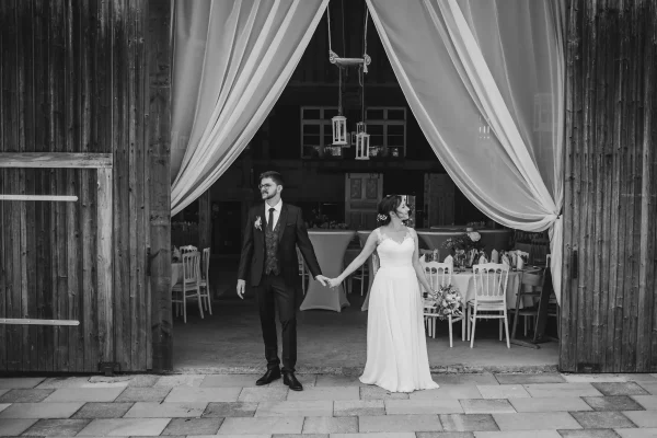 Das Brautpaar steht vor der rustikalen Scheune des Lindenhofs Mötzingen, umgeben von fließenden Stoffbahnen. Sie halten sich an den Händen und blicken voller Vorfreude in die Zukunft.