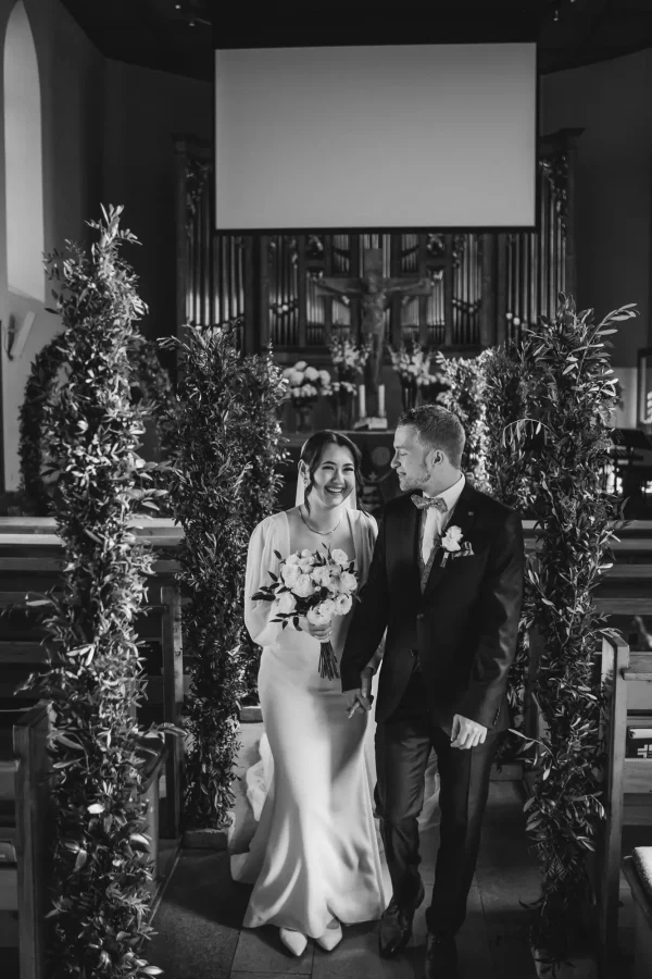 Das Brautpaar schreitet gemeinsam durch den mit Olivenzweigen geschmückten Mittelgang der Martinskirche. Der Bräutigam blickt seine Braut voller Liebe an, während sie mit strahlendem Lächeln ihren Brautstrauß hält.