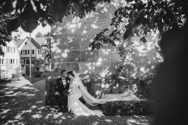 Das Brautpaar sitzt draußen auf einer Bank, umgeben von herbstlichem Lichtspiel und sanften Schatten der Blätter. Der lange Schleier der Braut fließt elegant über den Boden, während sie sich liebevoll anlehnen.