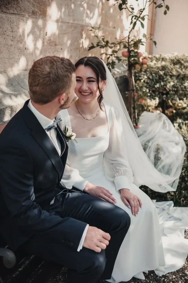Das Brautpaar sitzt auf einer Bank an der Kirchenmauer, während die Herbstsonne durch die Blätter schimmert. Die Braut strahlt ihren Bräutigam an, während ihr Schleier elegant im Hintergrund fließt.