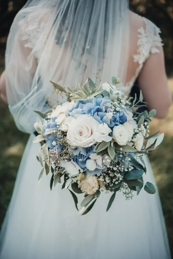 Detailaufnahme eines Brautstraußes in Blau-Weiß-Tönen, gehalten von der Braut mit fließendem Schleier – romantisches Hochzeitsdetail in sanftem Licht.