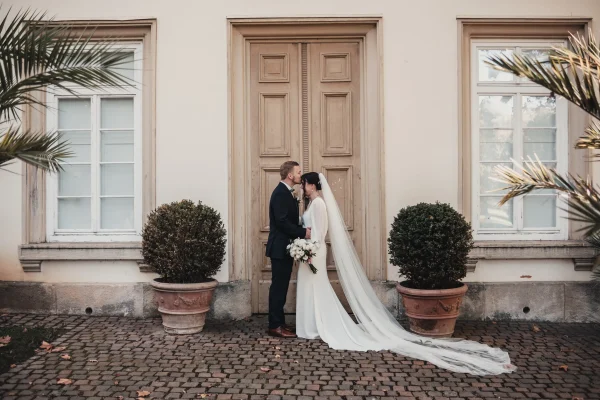 Romantisches Brautpaarshooting vor einer historischen Tür im Hohenheimer Park. Die Braut hält ihren Brautstrauß, während der Bräutigam ihr sanft die Stirn küsst. Mediterrane Elemente wie Palmen und Terrakotta-Töpfe sorgen für eine stilvolle Kulisse.