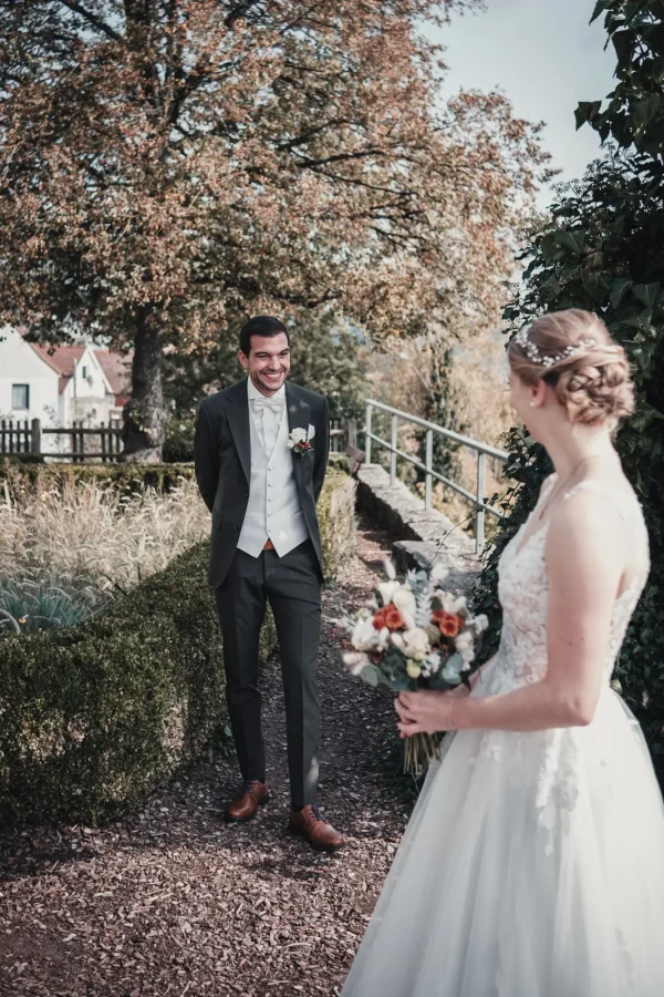 Der Bräutigam wartet lächelnd auf seine Braut im Garten der Burg Reichenberg. Die herbstliche Kulisse unterstreicht die Romantik des First Looks.”