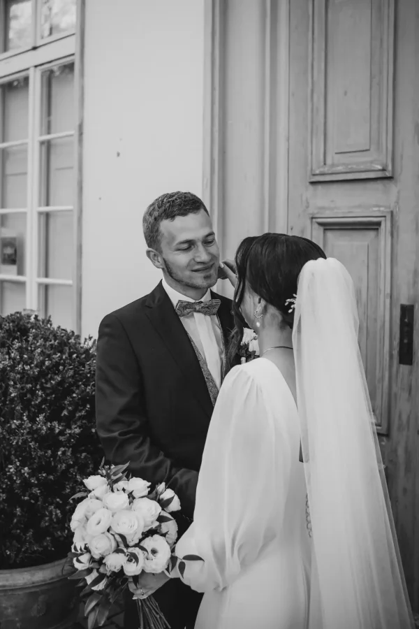 Ein emotionaler Moment vor einer historischen Tür: Die Braut lehnt sich liebevoll an den Bräutigam, ihr langer Schleier fließt elegant auf den Boden. Herbstliche Blätter und mediterrane Pflanzen rahmen die Szene.