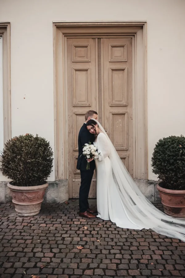 Romantische Kulisse im Hohenheimer Park: Das Brautpaar steht vor einer eleganten Holztür, ihre Stirnen berühren sich sanft. Der Brautstrauß mit weißen Rosen sorgt für einen klassischen, stilvollen Akzent.