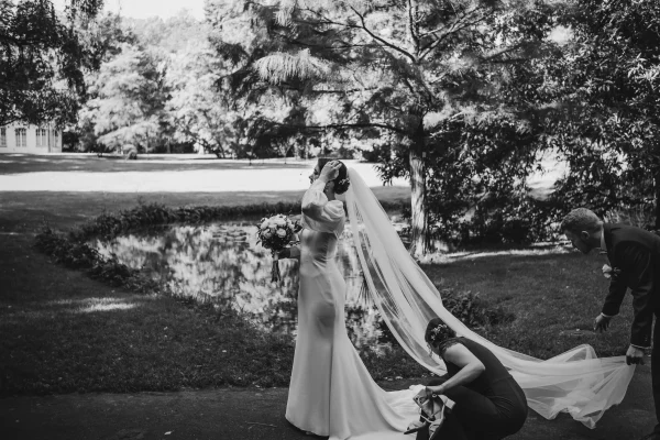 Ein stimmungsvoller Schwarz-Weiß-Moment: Die Braut wird von ihren Liebsten für das Shooting vorbereitet, während der Bräutigam sich um ihren Schleier kümmert. Im Hintergrund spiegelt sich die Natur im Wasser des Parks.