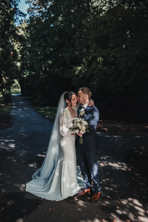 Das Brautpaar steht verliebt in einer von Bäumen gesäumten Allee. Die Braut hält ihren weißen Rosenstrauß, während sie sich liebevoll an den Bräutigam lehnt. Das Sonnenlicht zaubert ein wunderschönes Spiel aus Licht und Schatten.