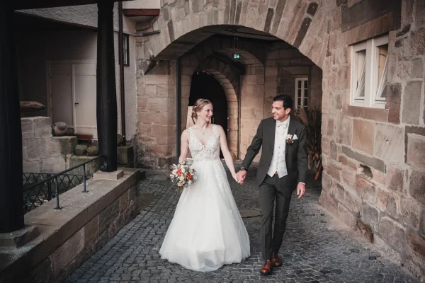 Das Brautpaar hält sich bei einem romantischen Spaziergang im Innenhof der Burg Reichenberg an den Händen. Die historische Architektur und das sanfte Licht im Hintergrund schaffen eine intime Atmosphäre.