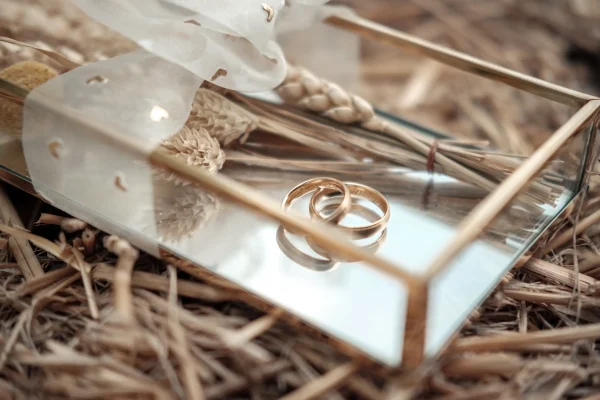 Zwei goldene Eheringe in einer eleganten Glasbox, dekoriert mit Schleierkraut und Weizen, auf einem Bett aus Stroh.