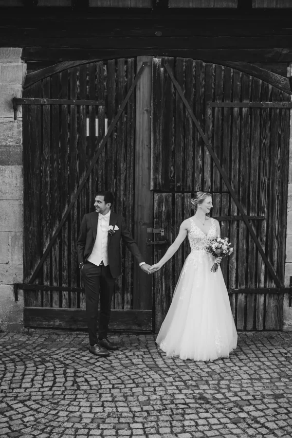 Das Brautpaar posiert mit einem Lächeln vor einer großen Holztür. Die Braut hält ihren wunderschönen Strauß und der Bräutigam hat einen charmanten Ausdruck auf dem Gesicht.