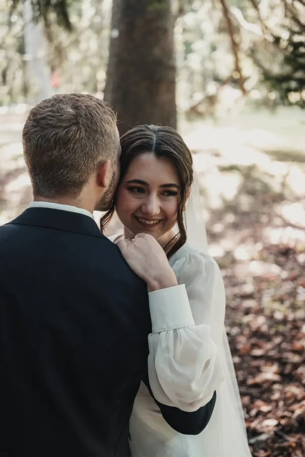 Innige Nahaufnahme: Die Braut hält sich am Bräutigam fest und blickt mit einem glücklichen Lächeln über seine Schulter. Die Sonnenstrahlen brechen durch das Laub und verleihen der Szene eine warme Atmosphäre.
