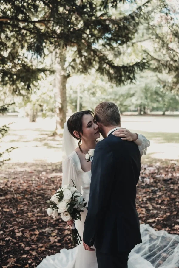 Emotionale Szene unter den Bäumen: Die Braut hält ihren Brautstrauß, während sie liebevoll den Bräutigam umarmt. Der lange Schleier fällt elegant über den Waldboden, während das Licht sanft durch die Äste schimmert.