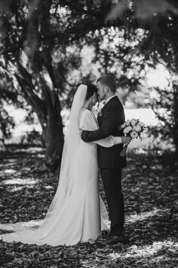 Schwarz-Weiß-Aufnahme eines romantischen Moments: Das Brautpaar steht inmitten eines Blätterwaldes, während der Bräutigam der Braut sanft die Stirn küsst. Der Brautstrauß aus weißen Rosen hebt sich kontrastreich ab.