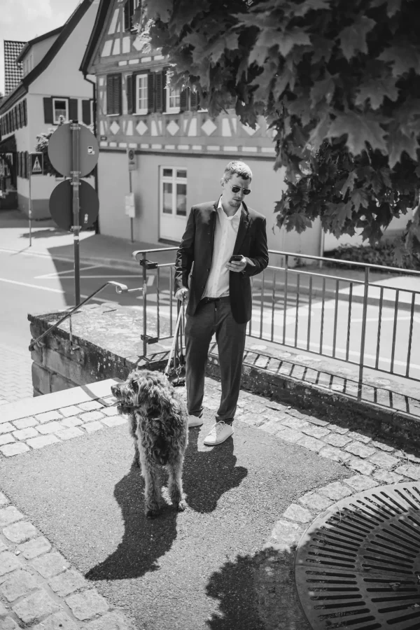 Eleganter Hochzeitsgast mit Sonnenbrille und Hund, stehend vor einer Fachwerkkulisse in Mötzingen.