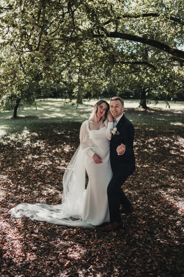 Verspielter Moment unter herbstlichen Bäumen: Der Bräutigam posiert mit einem Augenzwinkern, während die Braut sich lachend an ihn schmiegt. Ihr langer Schleier fällt sanft über das Laub.