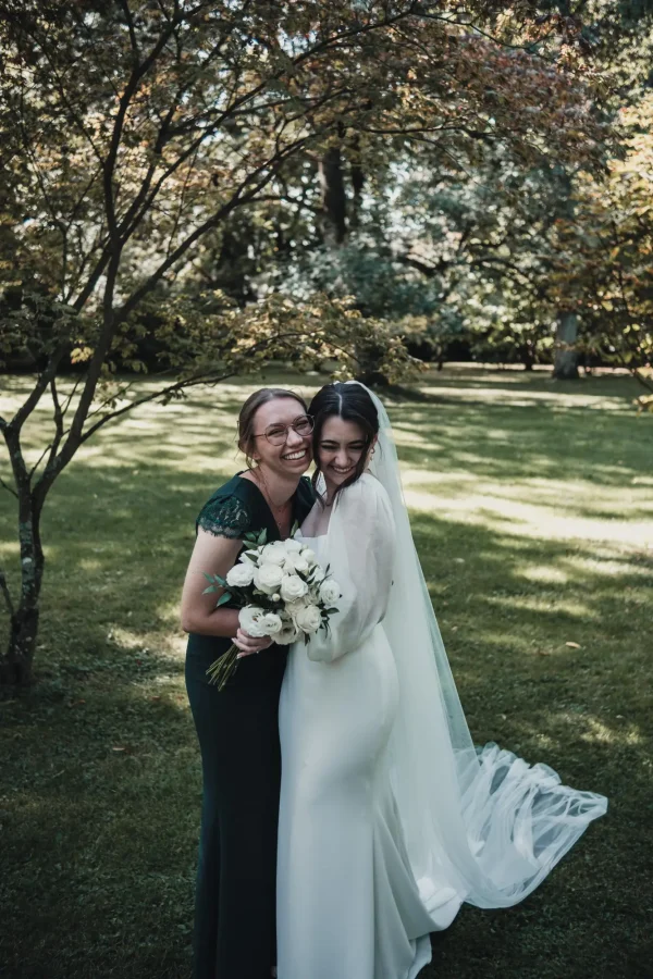 Die Braut umarmt eine enge Freundin lachend, während beide den Brautstrauß halten. Die herbstlichen Farben des Parks und das sanfte Sonnenlicht verleihen der Szene eine warme, fröhliche Atmosphäre.