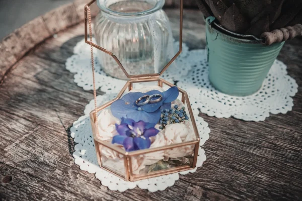 Detailaufnahme der Eheringe in einer eleganten Glasbox mit blauen Blüten – stilvolle Hochzeitsdekoration mit Vintage-Charme.