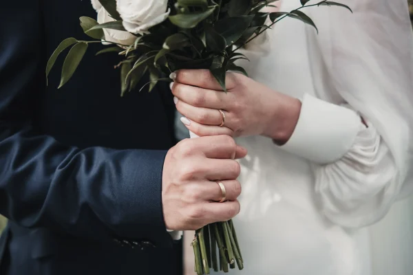 Symbolischer Moment der Verbundenheit: Das Brautpaar hält gemeinsam den Brautstrauß, während ihre Hände mit den glänzenden Eheringen sanft ineinander verschlungen sind.
