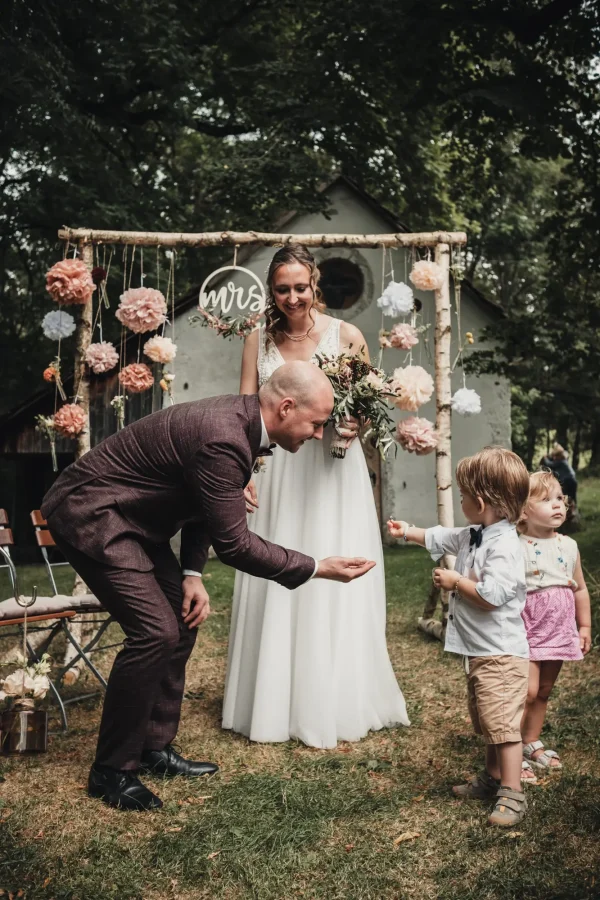Der Bräutigam beugt sich mit einem Lächeln zu zwei Kindern hinunter, während die Braut mit Blumenstrauß im Hintergrund strahlt. Die romantische Dekoration mit Papierblumen und einem “Mrs”-Schild rahmt die Szene.