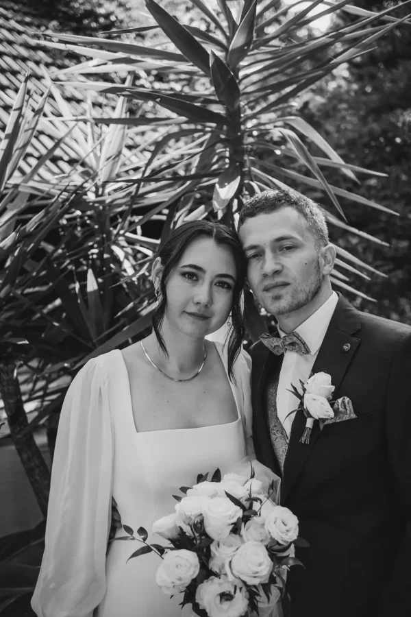 Schwarz-Weiß-Aufnahme mit mediterranem Flair: Das Brautpaar posiert vor großen Palmenblättern, während die Braut ihren weißen Rosenstrauß hält. Ein klassisches, stilvolles Portrait voller Eleganz.