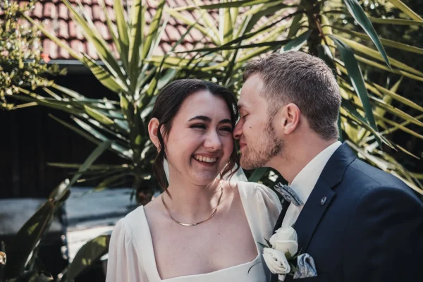 Glücklicher Moment voller Leichtigkeit: Die Braut lacht ausgelassen, während der Bräutigam ihr sanft einen Kuss auf die Wange gibt. Die exotische Kulisse mit großen Palmen verleiht der Szene eine besondere Note.