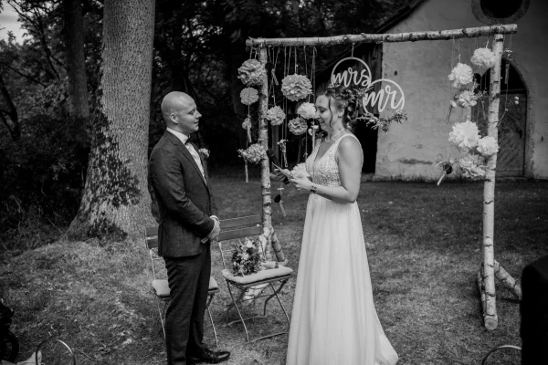 Emotionale Trauungsszene: Die Braut hält ein Mikrofon und trägt ihr Ehegelübde vor, während der Bräutigam mit liebevollem Blick zuhört. Die Kulisse mit rustikaler Hochzeitsdeko und Holzelementen hebt sich dezent ab. (Schwarz-Weiß-Bild)