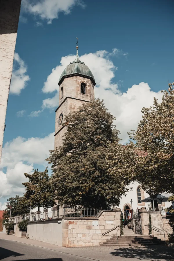 Die Martinskirche Sielmingen in herbstlicher Kulisse: Der historische Kirchturm ragt in den blauen Himmel, während Hochzeitsgäste sich vor dem Eingang versammeln. Eine idyllische Außenaufnahme der Trauungslocation.
