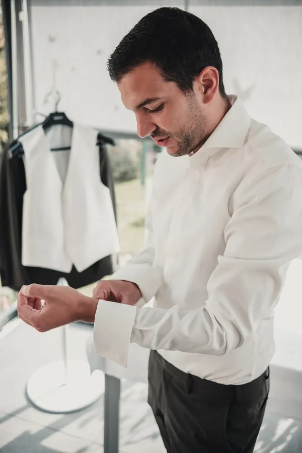 Bräutigam im weißen Hemd beim Anlegen der Manschettenknöpfe, festgehalten in einem lichtdurchfluteten Raum vor dem Anzug.
