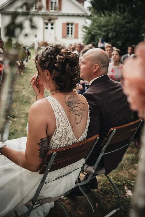 Braut und Bräutigam sitzen bei der freien Trauung auf rustikalen Holzstühlen, während die Braut gerührt eine Träne wegwischt. Im Hintergrund das malerische Hofgut Uhenfels mit Hochzeitsgästen.