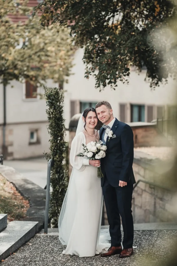 Strahlendes Brautpaar vor der Martinskirche Sielmingen: Die Braut hält ihren weißen Rosenstrauß, während sie und der Bräutigam lachend für ein Foto posieren. Herbstliche Blätter und eine geschmückte Treppe rahmen die Szene ein.