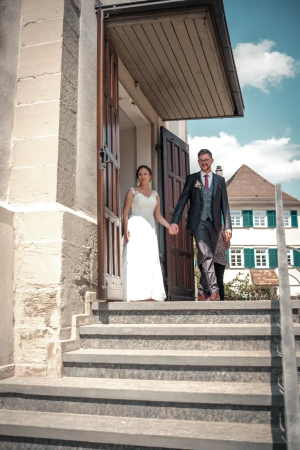 Das frisch vermählte Ehepaar Ena & Daniel verlässt Hand in Hand die Mauritiuskirche Mötzingen, strahlend unter blauem Himmel.