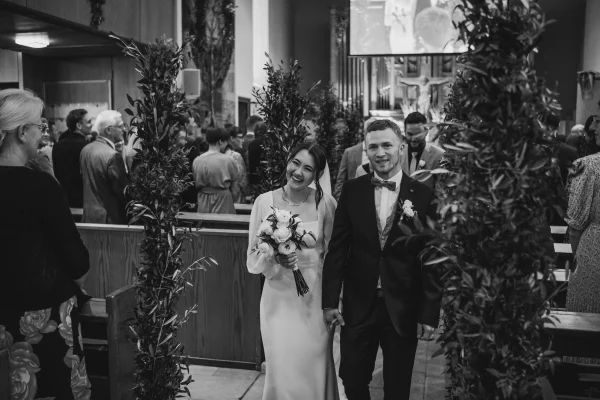 Schwarz-Weiß-Fotografie eines glücklichen Brautpaars beim Auszug aus der Kirche. Die Braut lächelt strahlend, während der Bräutigam ihre Hand hält. Gäste und Kirchenbänke im Hintergrund.