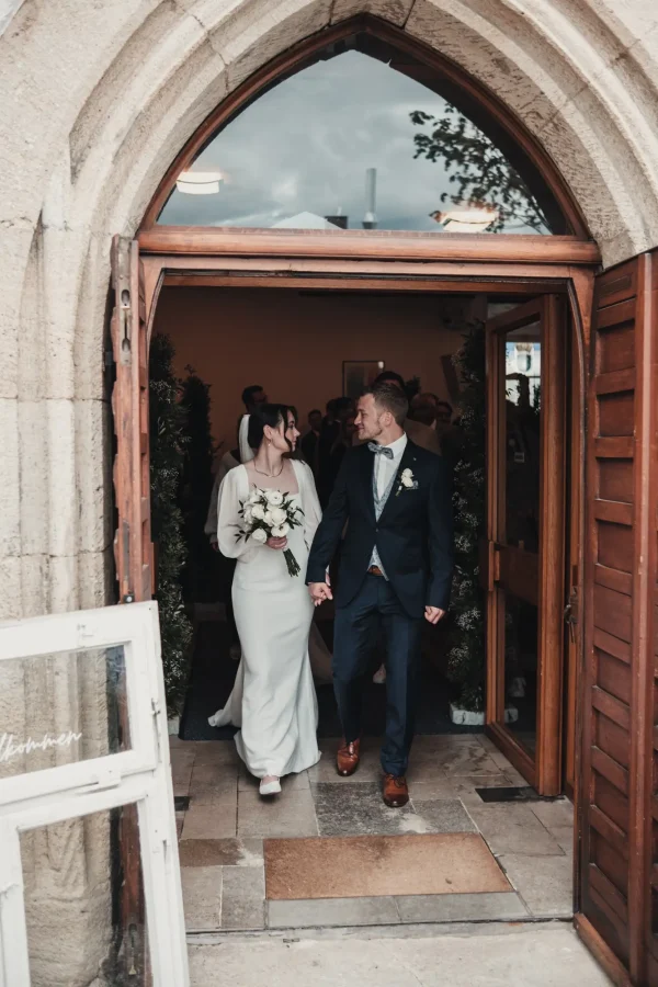 Frisch vermähltes Paar verlässt Hand in Hand die Kirche durch ein gotisches Portal, lächelnd und voller Freude. Die Braut hält einen Blumenstrauß mit weißen Rosen.