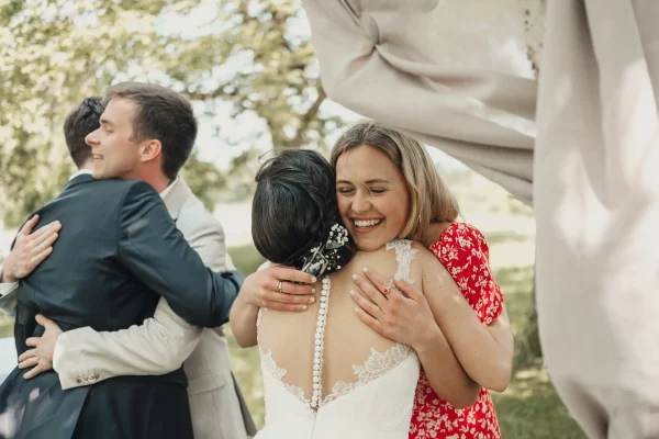 Die Braut wird strahlend von einer Freundin umarmt, während die Hochzeitsgäste im Hintergrund gratulieren. Ein emotionaler Moment voller Freude und Herzlichkeit im grünen Außenbereich der Hochzeitslocation.