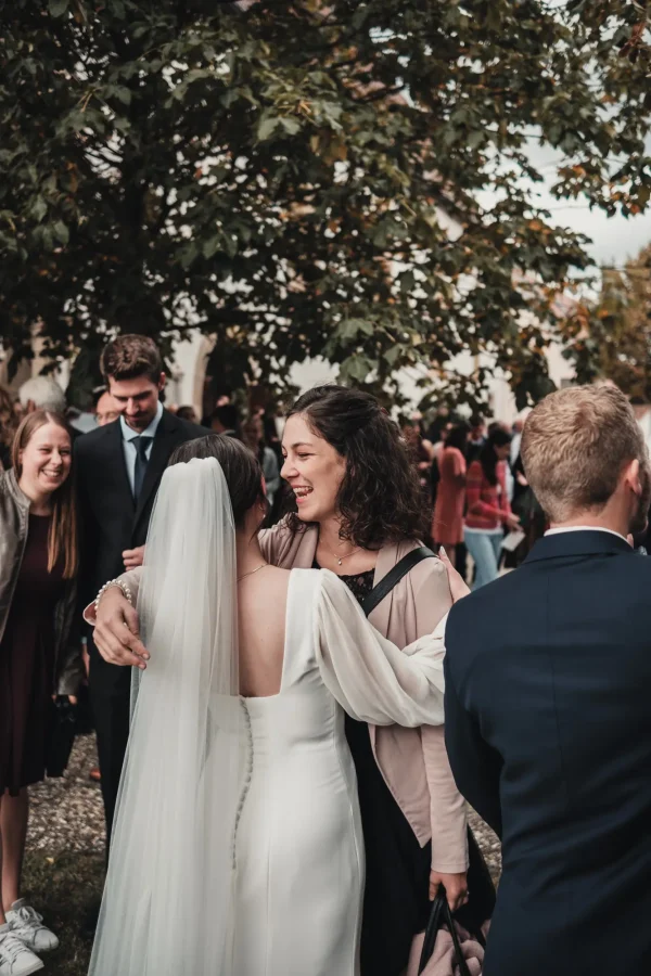 Die Braut umarmt eine glückliche Hochzeitsgästin nach der Trauung. Im Hintergrund stehen lachende Gäste unter einem großen Baum.