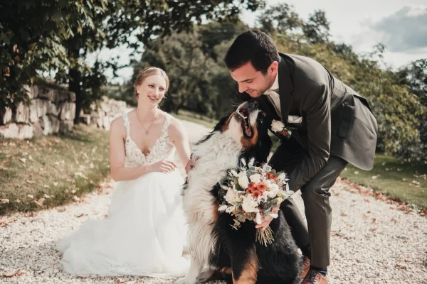 Der Bräutigam hält einen Blumenstrauß, während er liebevoll mit dem Berner Sennenhund posiert, die Braut strahlt im Hintergrund.