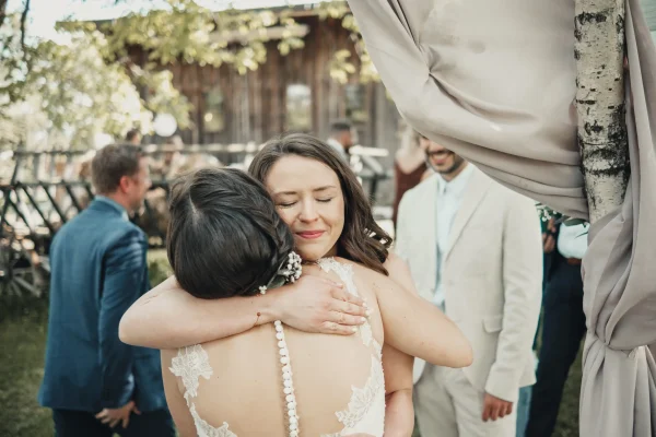 Die Braut wird liebevoll von einer Freundin umarmt, die mit geschlossenen Augen den Moment genießt. Im Hintergrund feiern Gäste inmitten der ländlichen Hochzeitslocation Lindenhof Mötzingen.
