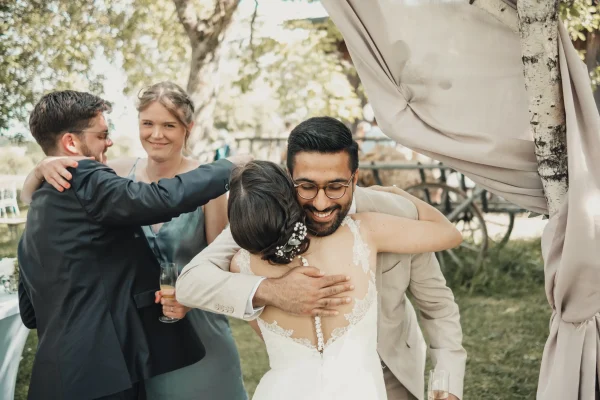 Die Braut umarmt freudig einen Hochzeitsgast, während sich der Bräutigam mit einer weiteren Person austauscht. Ein bewegender Moment voller Herzlichkeit und Freude in der rustikalen Hochzeitslocation Lindenhof Mötzingen.