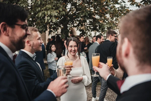 Die Braut lacht strahlend während des Sektempfangs, umgeben von Gästen, die mit Sekt und Orangensaft anstoßen. Eine fröhliche Atmosphäre unter Bäumen und festlich gekleideten Freunden.
