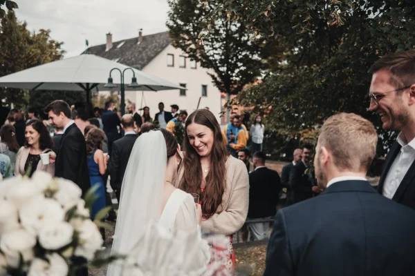 Die Braut spricht mit einer lachenden Hochzeitsgästin während des Sektempfangs. Im Vordergrund ist ein Brautstrauß mit weißen Rosen sichtbar, während sich Gäste im Hintergrund unterhalten.