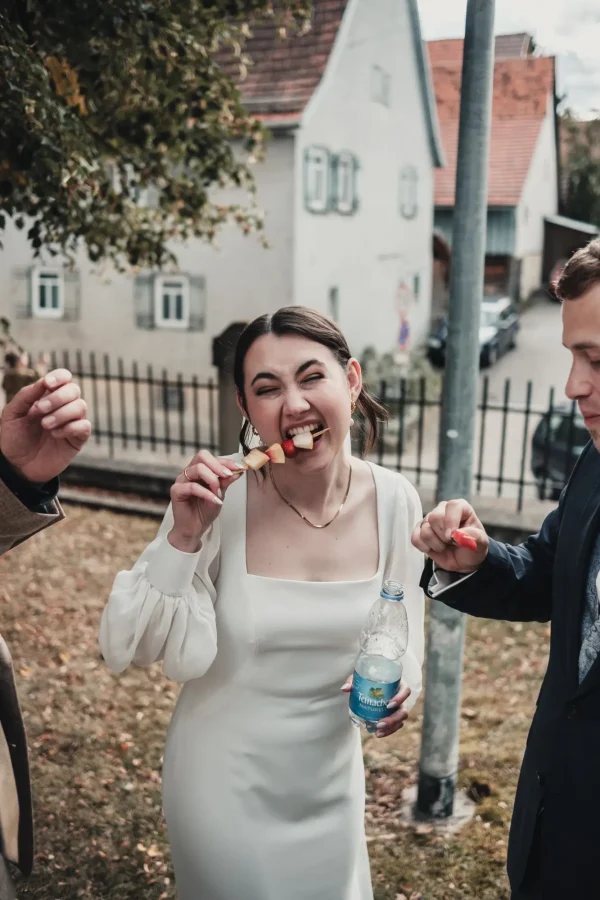 Die Braut beißt mit einem fröhlichen Gesichtsausdruck in einen Obstspieß, während sie eine Wasserflasche hält. Neben ihr stehen weitere Hochzeitsgäste, die sich am Essen erfreuen.