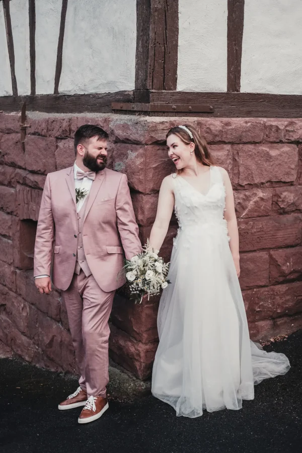Brautpaar lacht sich herzlich an im Außenbereich der Pfrondorfer Mühle. Steinwand und Fachwerk im Hintergrund