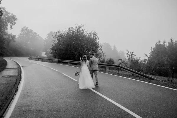 Brautpaar spaziert Hand in Hand auf einer nebligen Landstraße, eingefangen in einer stimmungsvollen Schwarz-Weiß-Aufnahme